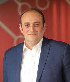 A middle-aged, fair skinned with dark hair partially covering his hair, man smiling towards the camera. 