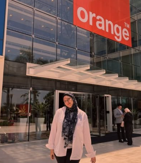 A veiled female is standing in front of a building. There is a hanging sign with the text: Orange